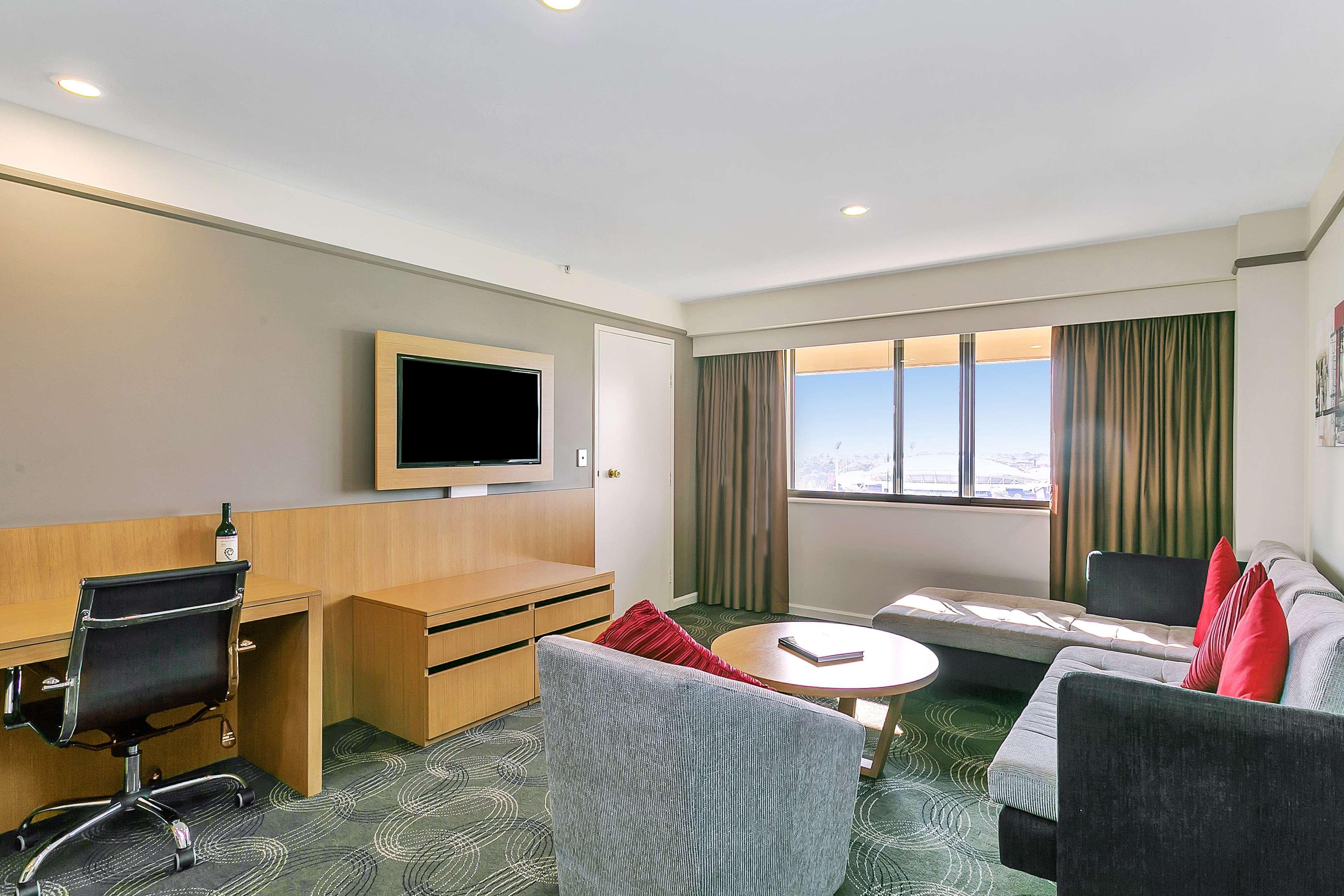 Stamford Plaza Adelaide Hotell Eksteriør bilde The photo shows a modern hotel room or suite that features a cozy seating area. There is a comfortable sofa with red accent pillows, a round coffee table in the center, and a sleek desk with an office chair. On the wall, there is a flat-screen TV mou